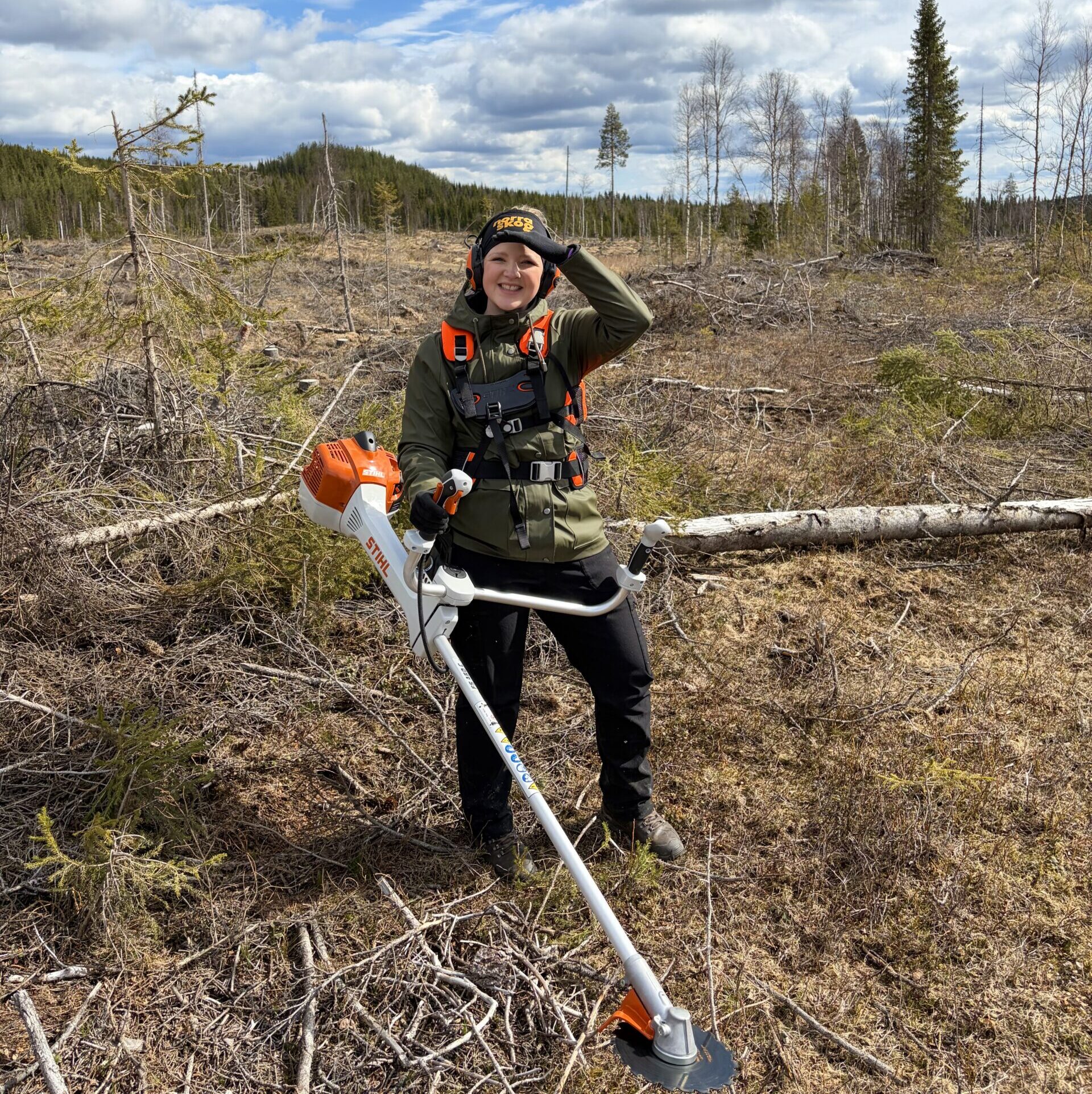 Julia står i skogen och har på sig röjsågsselen. Det är soligt väder.