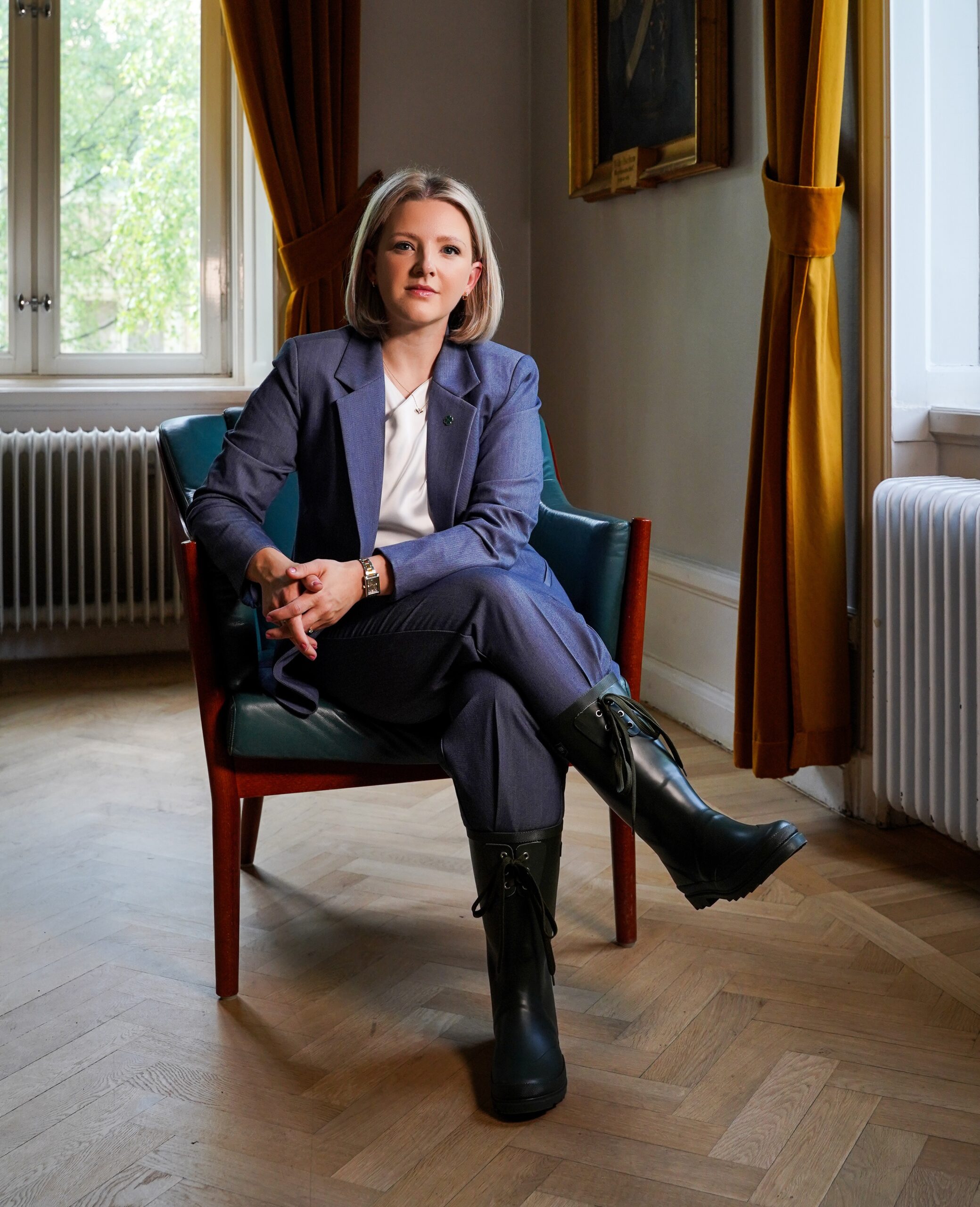 Julia Algotsson, iklädd blå kostym med gröna stövlar. Sitter i en fåtölj.